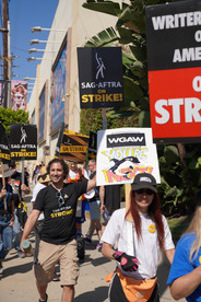 A candid shot of protesters. Most visible in the photo is a man and woman. The man wears a "SAG-AFTRA Strong" t-shirt with his protest sign raised high abovehead; the woman wears a black WGA baseball cap and holds a sign that says "AMPTP You're Next!" with a drawing of an orca whale.