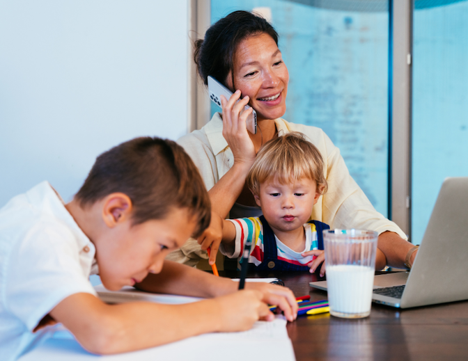 A mother multitasking at home, talking on a smartphone and using a laptop while helping her two young children with schoolwork, representing "time poverty" and caregiving burdens.