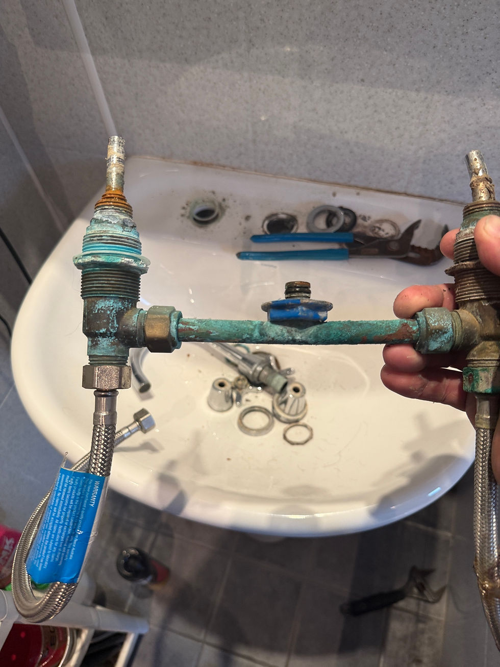 Close-up of a heavily corroded basin tap assembly with green oxidation on copper components, removed and held above a bathroom sink during repair.