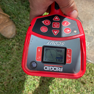 Hand holding a pipe locator device displaying readings, used to identify the position of an underground stormwater drain.