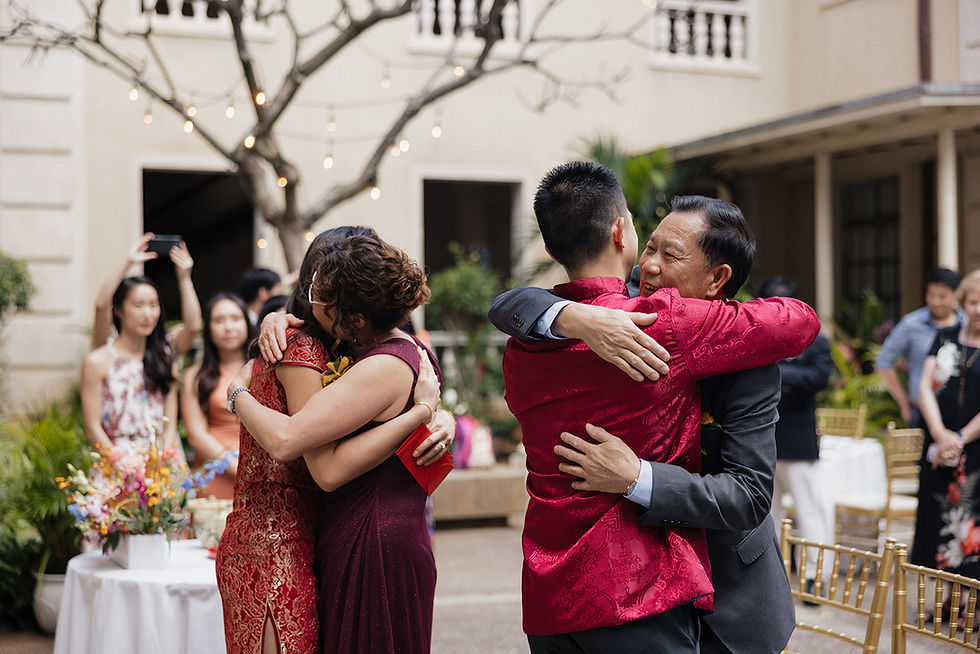 Four Chinese heritage people hug joyfully in a garden with string lights and chairs. Two wear red outfits. Onlookers and potted plants are in the background.