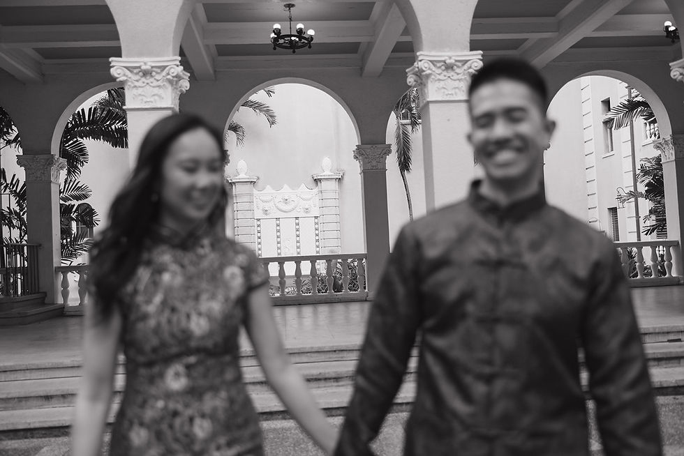 Blurred image of a smiling Chinese couple in traditional clothing holding hands at wedding venue. They stand in front of ornate arches with Hawaiian palm trees in the background.
