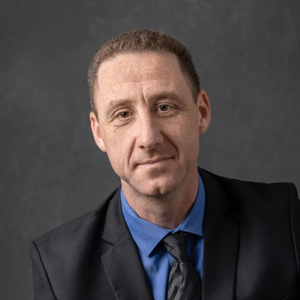 תמונה של גבר לבוש בחליפה כהה, חולצה כחולה ועניבה, רקע אפור כהה