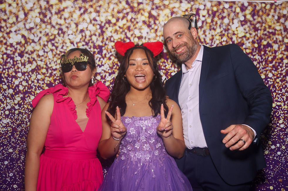 Three people pose in colorful outfits; one woman wears "Congrats" glasses, another shows peace signs. Glittery purple backdrop adds festivity.