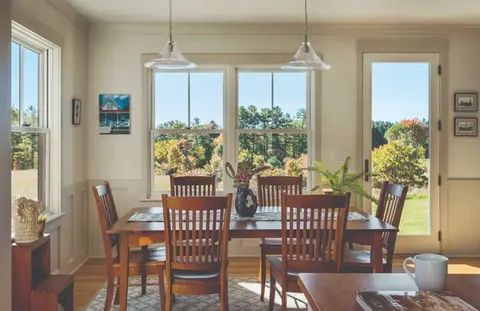 Bright dining room with wooden table, chairs, and large windows offering scenic views