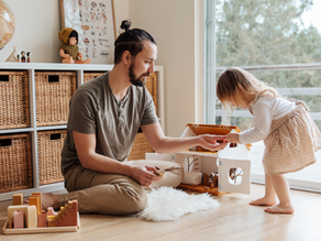 Un père joue avec sa fille en bas âge dans un salon lumineux, entouré de jeux éducatifs et de rangements en rotin. Un moment de complicité parent-enfant illustrant l’implication des pères dans la parentalité et le quotidien familial.