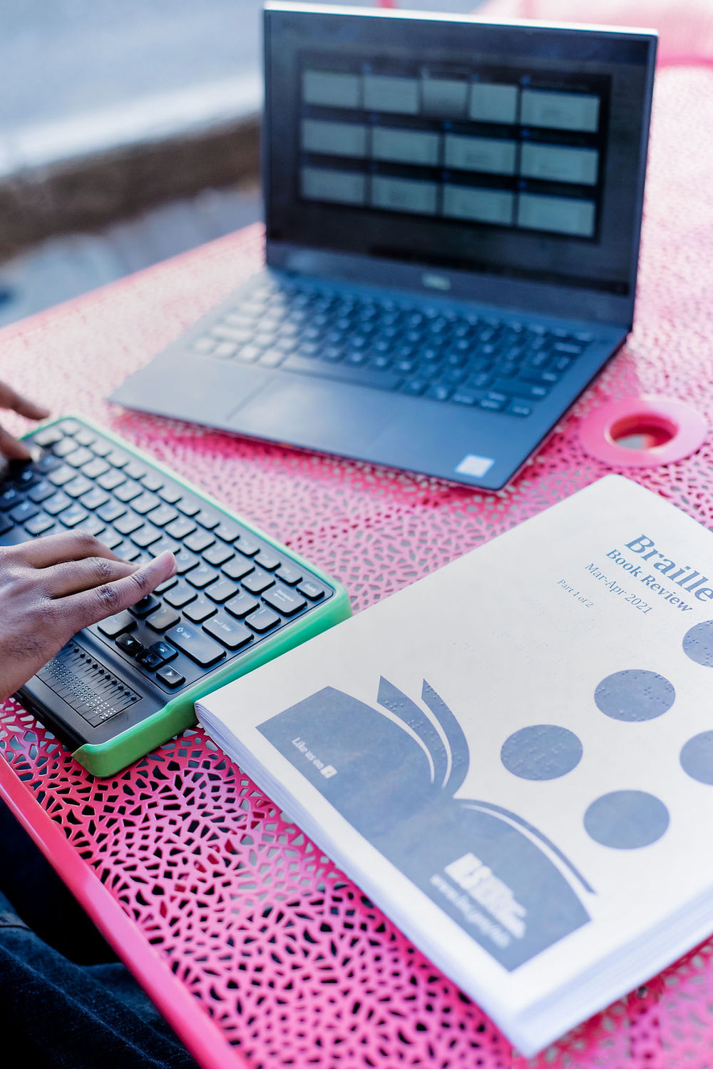 A table that has a Dell laptop showing the Windows Task Switcher screen, a closed Braille Book Review, and hands typing on the Mantis Braille Display