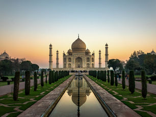 The Taj Mahal mausoleum is captured in a perfectly symmetrical shot during sunrise or sunset. The warm light of the sky casts a golden glow on the white marble structure. In the foreground, the iconic reflecting pool leads the viewer's eye towards the monument, flanked on both sides by lush green lawns and dark cypress trees.