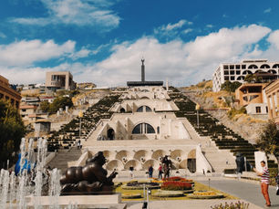 The Cascade Complex in Yerevan, Armenia, a monumental white limestone staircase built into a hillside, with fountains and modern art sculptures at its base. People are strolling around the complex under a bright blue sky on a clear day