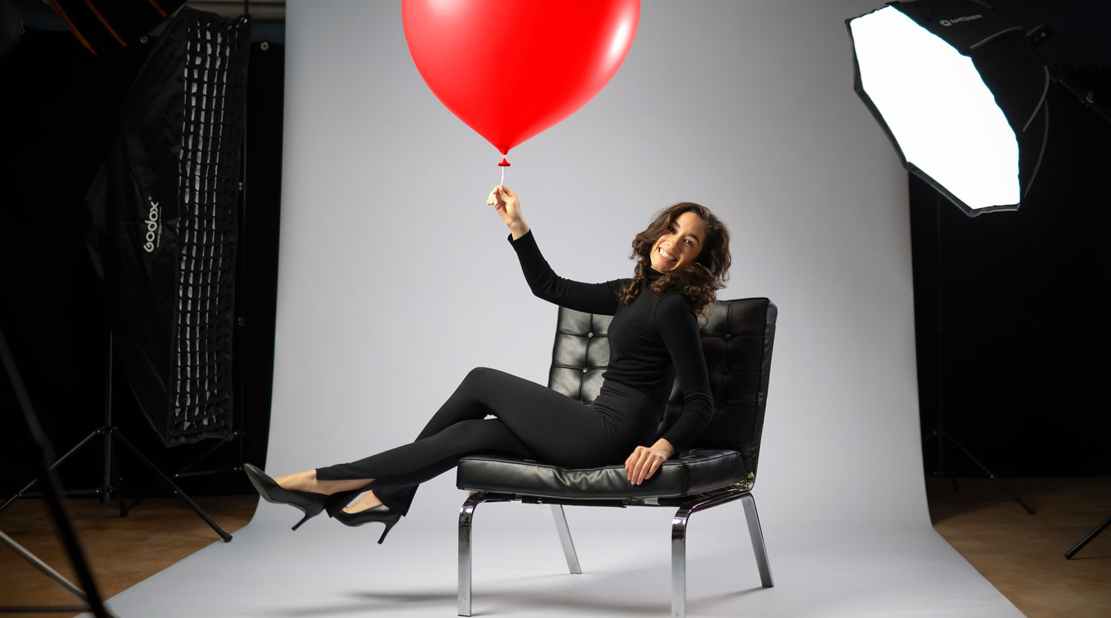 Model im Studio während des Shootings sitzend in schwarz gekleidet und rotem Luftballon in der Hand