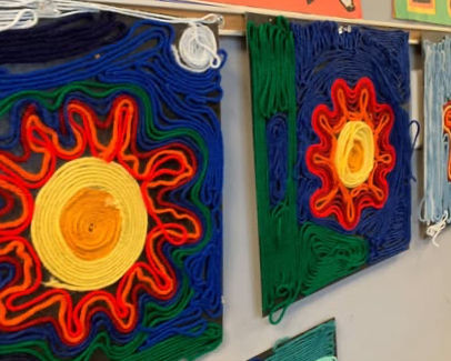 Colorful yarn wall art with abstract flower designs in blue, red, green, and yellow hanging in a classroom.