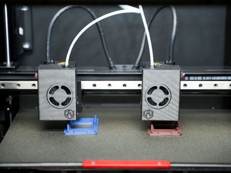 Raise3D E3 em IDEX: dois extrusores imprimindo peças simultâneas (azul e vermelho) sobre mesa PEI