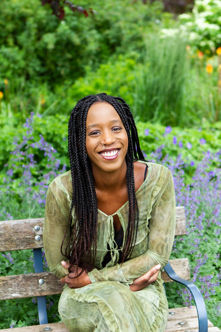 outdoor image of black girl in a gardens sitting on bench with a huge smile on her face, almost laughing into the pose. the black girl is wearing green see through dress complimenting the background. Girl has brown skin and long black box braids 