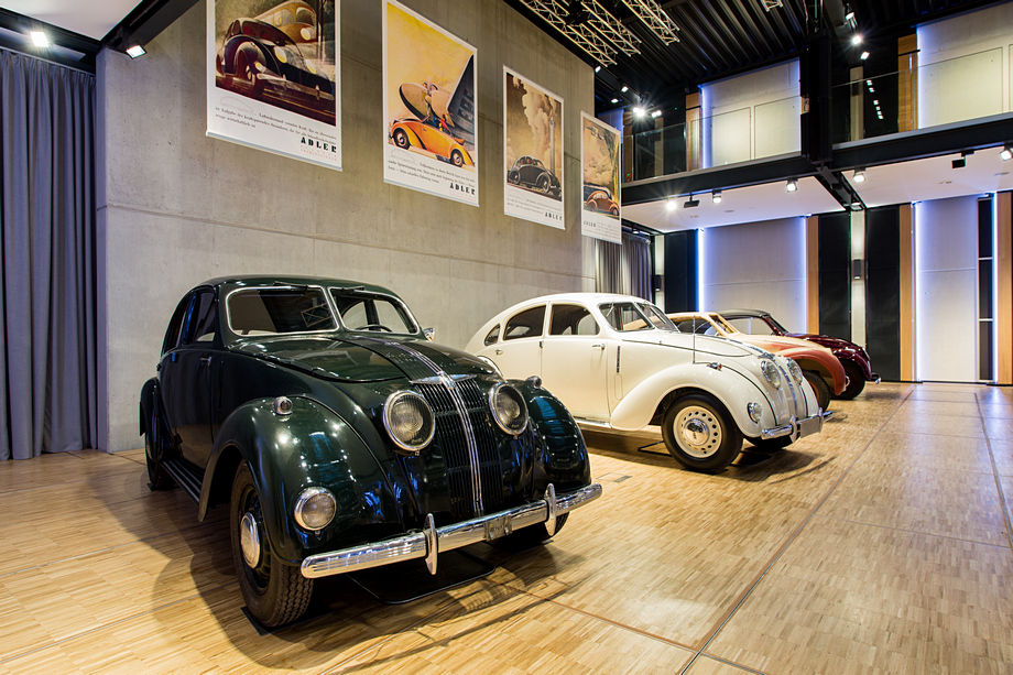 Drei stromlinienförmige Oldtimer der Vorkriegszeit stehen nebeneinander auf Parkettboden in einer modernen Ausstellungshalle mit Werbepostern an der Wand.