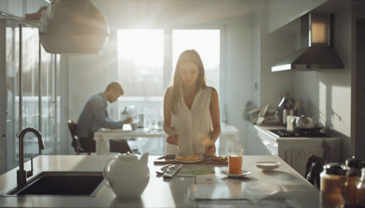 Mulher prepara o café da manhã enquanto o parceiro permanece distante ao fundo, representando o desequilíbrio no relacionamento e a sobrecarga emocional.