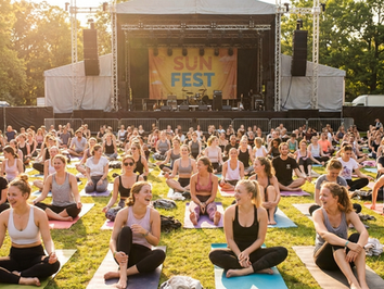 A large group of young adults doing yoga in a sunny park with a concert stage in the background