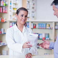 A bright photo of a pharmacist handing a small paper bag to a patient near a community park.