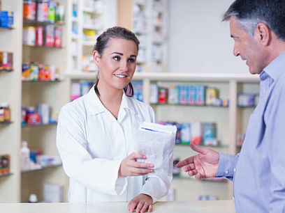 A bright photo of a pharmacist handing a small paper bag to a patient near a community park.