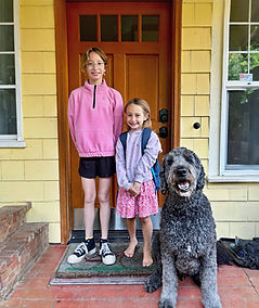 Two girls with backpack, happy family dog on yellow front porch.
