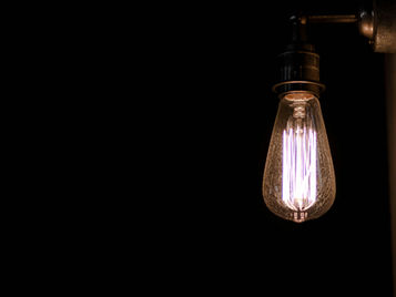 Lit vintage Edison bulb hanging against a dark wall, glowing warm white with a copper fixture