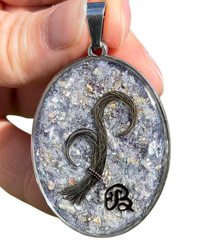 Photo of a pendant in silver stainless steel. Dog hair worked in 2 loops, attached by a beaded link. Dog ashes enclosed in resin, in a mixed purple background. Initial B written in dark ink.