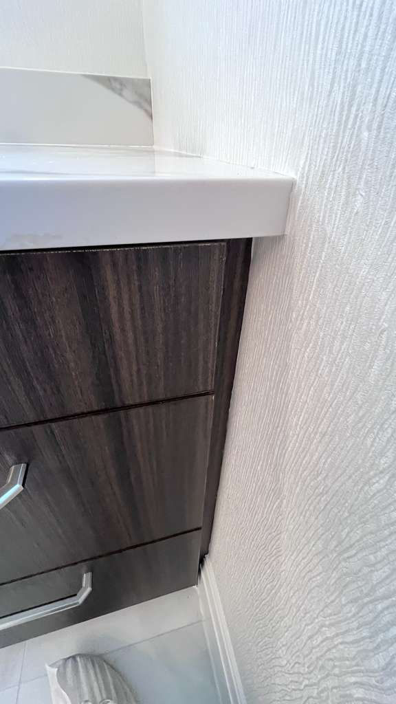 Dark wood cabinet beside a textured white wall in a tiled room. The countertop is white with a subtle pattern. A striped item is on the floor.