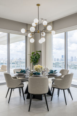 Contemporary dining room with a black and gold multi-bulb chandelier, a round table with a dark base, and beige dining chairs. Sheer curtains hang beside large windows showcasing an urban landscape.