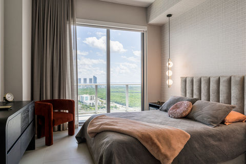 Elegant bedside arrangement in a contemporary bedroom with a white nightstand, a glass-topped surface, a hanging teardrop-shaped light, and a glimpse of a neatly made bed with a plush headboard and black out sheers