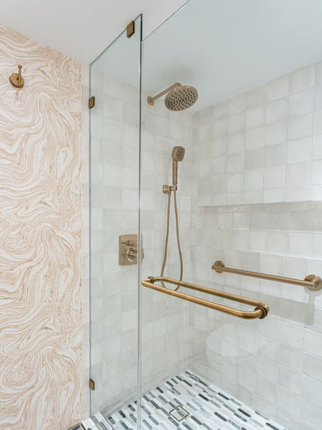 Modern shower with gold fixtures, a square rain showerhead, and a handheld spray. Beige marbled wall, white tiles, gray stripe floor.