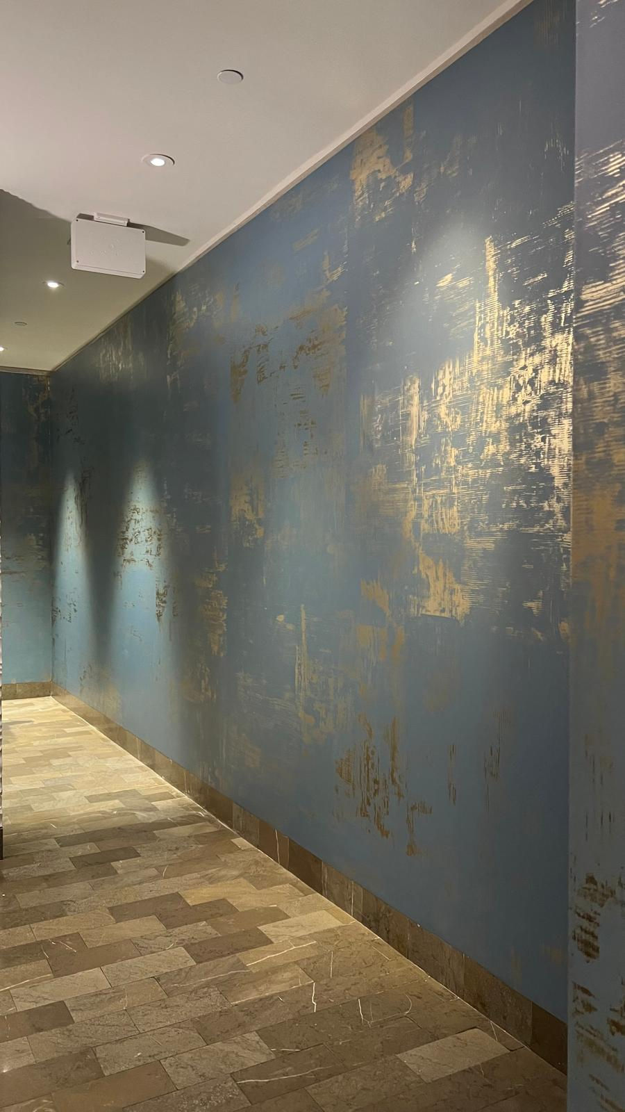 Hallway with textured blue and gold walls. Warm lighting casts shadows on the walls, and the floor is tiled in shades of beige and brown.