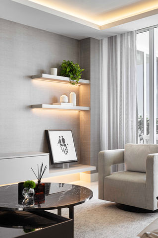 Interior view of a contemporary living space with a focus on a textured gray wall with two floating shelves illuminated from below, showcasing minimalist decor and greenery. A framed artwork rests on a lower shelf. A round coffee table with glass and dark surfaces is in the foreground, and a neutral armchair is visible to the right, near sheer window coverings.