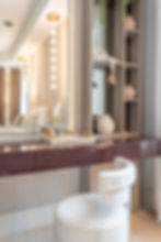 Elegant vanity with plush white chair, maroon tabletop, mirror, and shelves holding decor. Hanging lights create a warm ambiance.