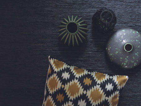 Geometric pillow with tan and black pattern alongside three textured vases on a dark wooden surface. Mood is calm and artistic.