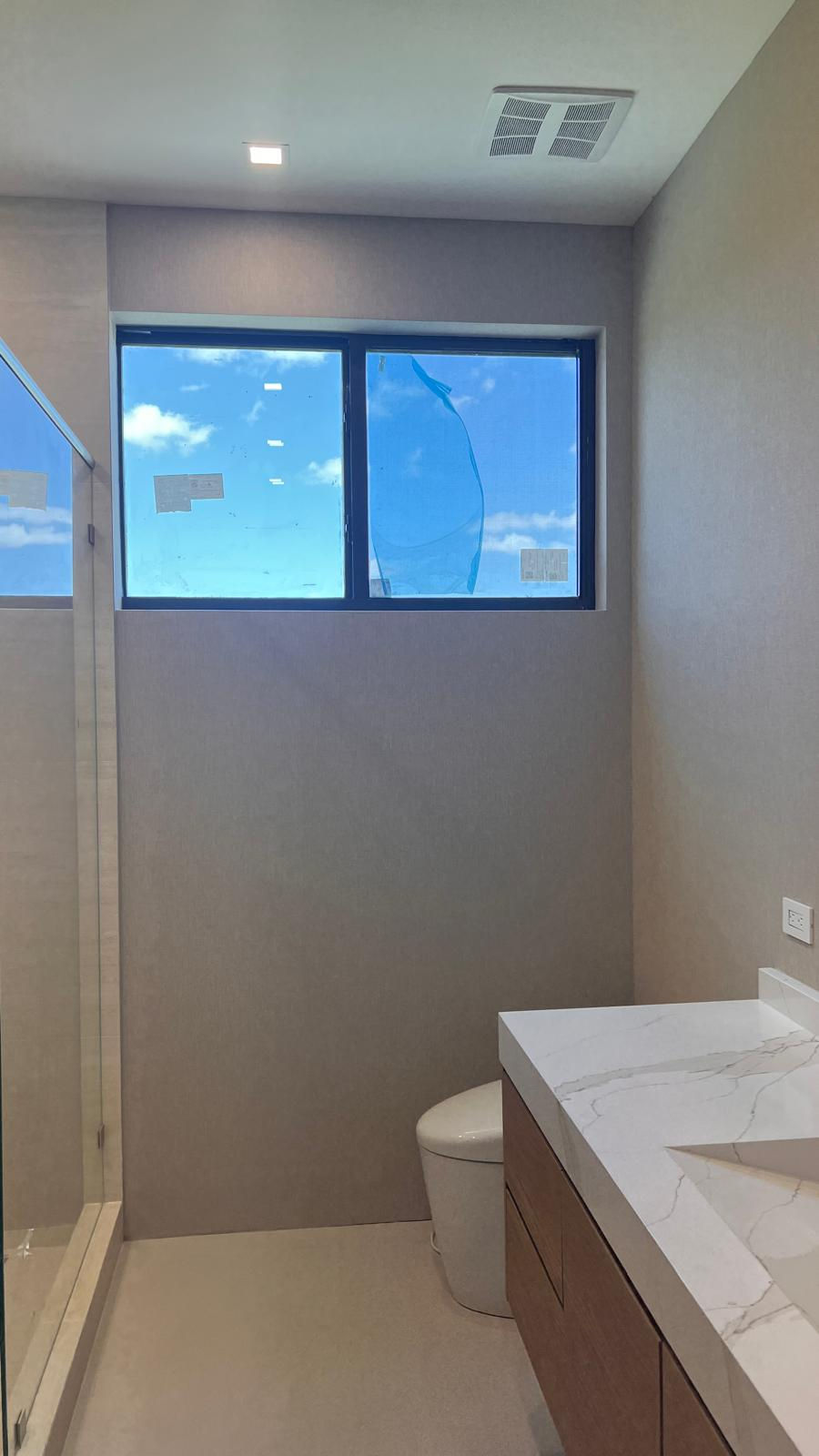 Modern bathroom with a large window showing a blue sky, a glass shower, white countertop, and a toilet. Neutral tones and bright light.
