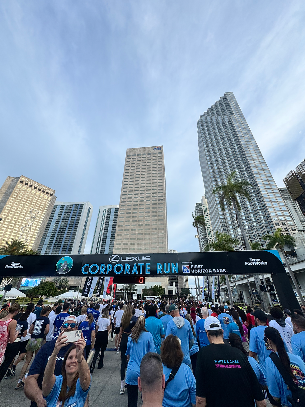 Crowd in blue shirts at Corporate Run under city skyscrapers, a banner overhead, with a clear sky. People are taking photos and smiling.