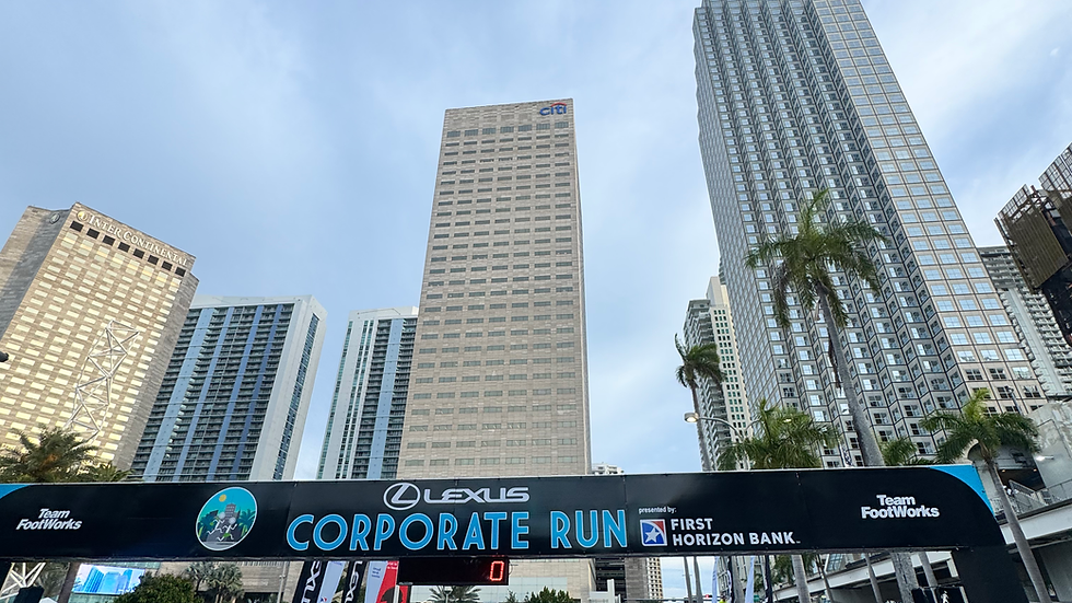 Crowd in blue shirts at Corporate Run under city skyscrapers, a banner overhead, with a clear sky. People are taking photos and smiling.