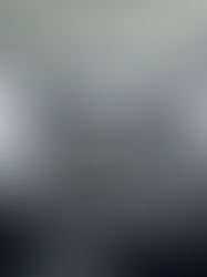 Home office with a wooden desk, black chair, and closed shutters. A round white table with books sits on a gray rug. White walls and minimal decor.