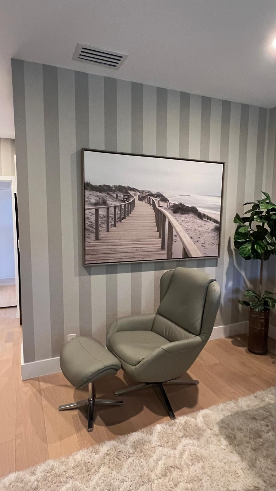Green chair and ottoman in a cozy room with a striped wall. Beach boardwalk painting above and a potted plant nearby on wooden floor.