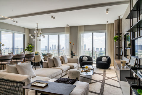 A neutrally colored living room in Miami, Florida, featuring a textured gray wall with two lit shelves holding decorative objects and a plant. A framed piece of art leans on a lower shelf. A round coffee table with a reflective surface is in the foreground, and a comfortable-looking beige armchair sits near sheer curtains and a window showing outdoor light.