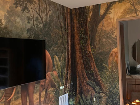 Corner angle of a Pinecrest playroom featuring a custom jungle safari wall mural, showing large ancient trees, travertine tile floors, and precise trimming around an en-suite bathroom doorway.