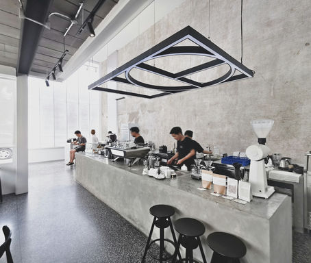 Modern café with staff behind a concrete counter, preparing drinks. Minimalist design, large windows, black stools, and ceiling lights.
