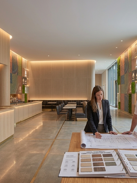 A wide-angle architectural photograph capturing a fully realized luxury hospitality lobby that unifies multiple wall surfaces. On the left, a curved reception desk is finished in fluted Natural Rift White Oak wood veneer. In the center, a micro-perforated acoustic felt wall accent framing leather booth seating provides sound dampening. On the right, a multi-paneled mural and living moss wall line a corridor with floor-to-ceiling glass windows overlooking a garden. In the foreground, an interior designer and an installer review architectural blueprints and an open sample binder titled 'Dress Your Walls: Complete Project Solutions' on a wooden table, illustrating end-to-end service from specification to final installation.