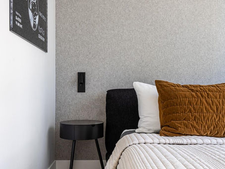 Cozy bedroom with gray walls, black side table, and a bed with a brown pillow. A wall art features "American Dream: Take Risks". Peaceful ambiance.