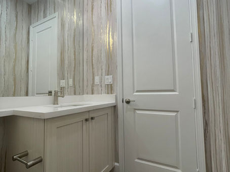 Bathroom with wood-patterned wallpaper, white vanity, and countertop. Wall lights reflected in a mirror, and a closed white door.