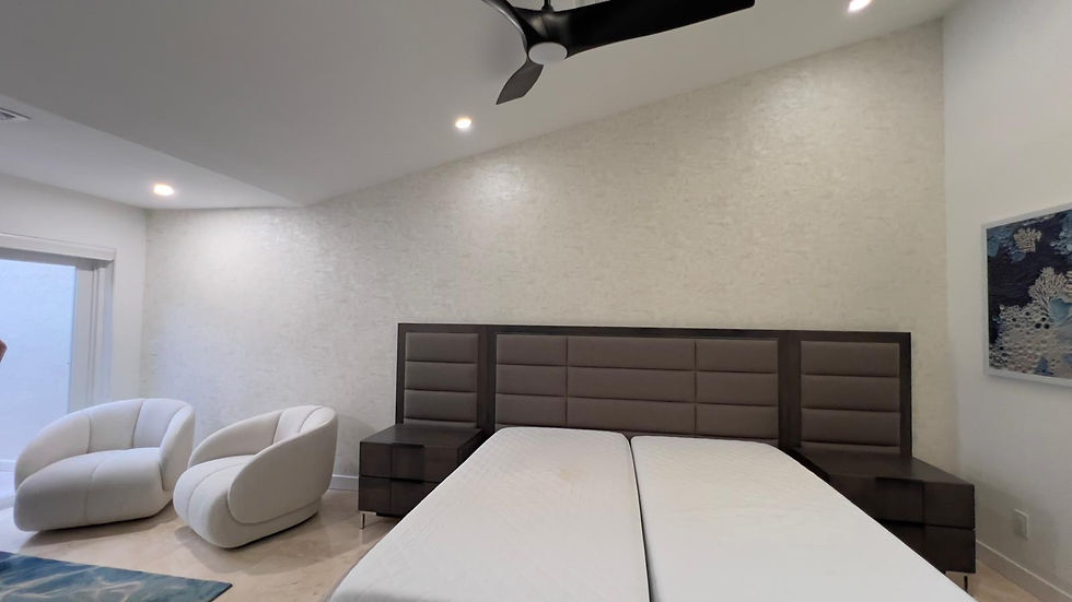 Modern bedroom with a large bed frame, two white chairs, and a ceiling fan. Light beige walls, blue rug, and abstract artwork on the wall.