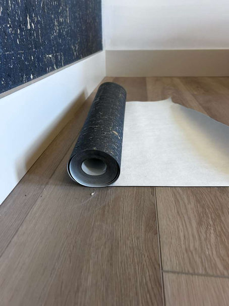 A roll of dark textured wallpaper on a light wooden floor, partially unrolled near a white baseboard. The setting is calm and minimalistic.