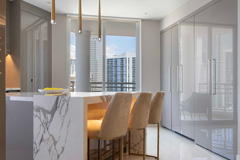 A bright and modern kitchen in a high-rise in Brickell, Florida, features a prominent white marble island with seating for three on beige and gold bar stools. Sleek gray cabinets line the wall, and pendant lighting illuminates the island. Large windows offer a view of the surrounding urban environment, possibly including the high-rise buildings of Brickell Key.