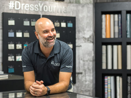 Man smiling in a wallpaper store with patterned rolls and samples. Text: "#DressYourWall." Wearing a gray shirt, cheerful atmosphere.