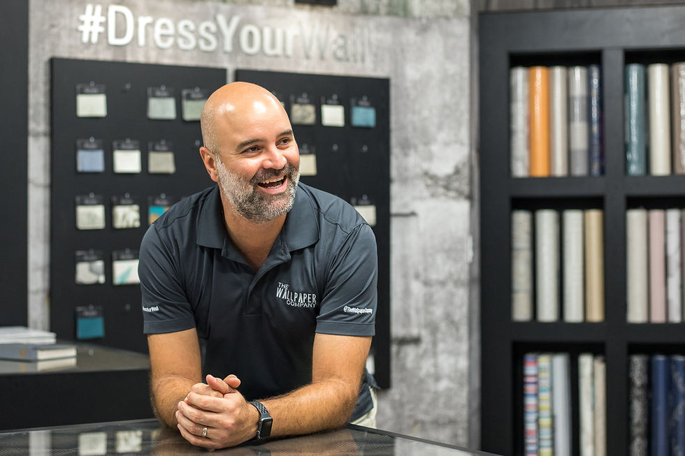 Man smiling in a wallpaper store with patterned rolls and samples. Text: "#DressYourWall." Wearing a gray shirt, cheerful atmosphere.
