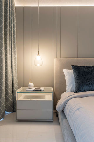 Detail of a modern bedroom interior showing a bedside table with a glass top, a hanging light fixture, a portion of a bed with a light headboard and neutral bedding, and patterned sheer curtains.